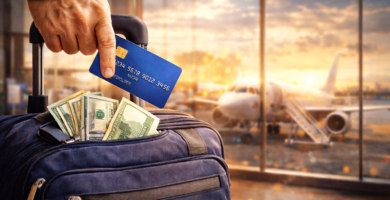 Mano sosteniendo una tarjeta de crédito frente a una maleta con billetes visibles, en un aeropuerto al atardecer con un avión en segundo plano.