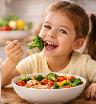 Niña sonriente disfrutando un plato de verduras frescas con brócoli, tomates cherry y pollo, en una cocina iluminada y acogedora.