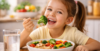 Niña sonriente disfrutando un plato de verduras frescas con brócoli, tomates cherry y pollo, en una cocina iluminada y acogedora.