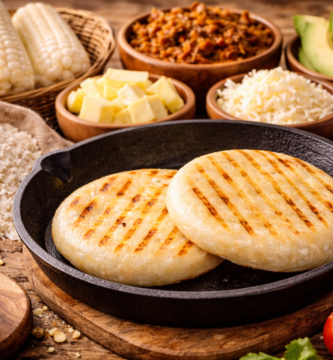 Primer plano de dos arepas doradas con marcas de parrilla en sartén de hierro, rodeadas de ingredientes como queso, aguacate, carne desmechada y salsa sobre mesa rústica.