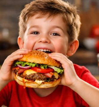 Niño sonriente de unos 7 años dando un gran mordisco a una hamburguesa casera con queso, bacon y vegetales frescos, acompañado de papas fritas y refresco al fondo