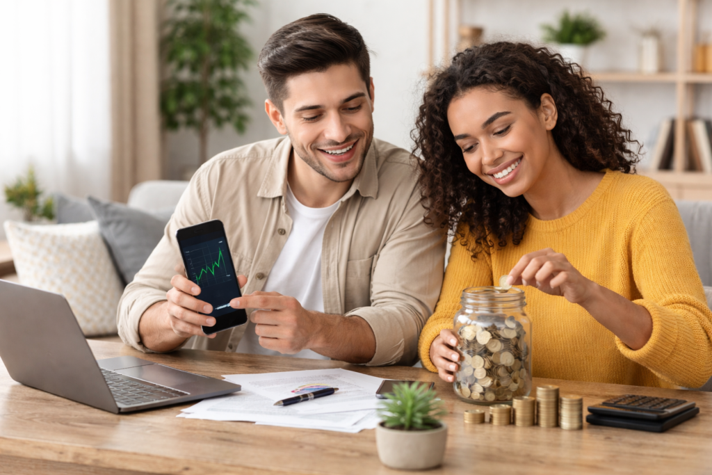 Pareja joven revisando inversiones en su móvil mientras ahorran dinero en un tarro en una mesa de trabajo, simbolizando cómo empezar a invertir desde cero.