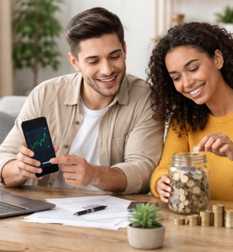 Pareja joven revisando inversiones en su móvil mientras ahorran dinero en un tarro en una mesa de trabajo, simbolizando cómo empezar a invertir desde cero.