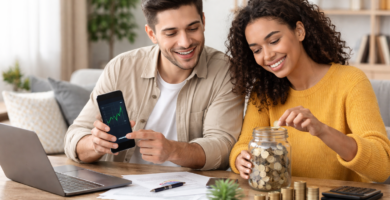 Pareja joven revisando inversiones en su móvil mientras ahorran dinero en un tarro en una mesa de trabajo, simbolizando cómo empezar a invertir desde cero.