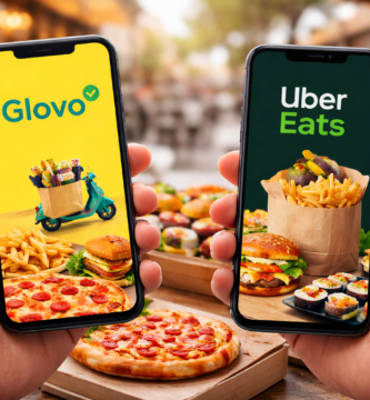 Dos teléfonos móviles mostrando las apps de delivery Glovo y Uber Eats con pizzas, hamburguesas, sushi y papas fritas al fondo en una mesa de comida al aire libre