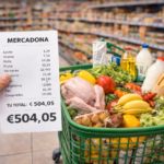 Carrito de supermercado lleno con productos básicos como pollo, leche, frutas, verduras, huevos y pan, junto a una mano que sostiene un ticket de compra de Mercadona por valor de 504,05 € en un pasillo de supermercado en España en 2026