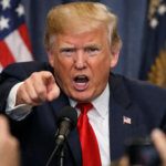 Donald Trump señalando de forma enérgica hacia el frente durante una rueda de prensa, con gesto serio y boca abierta mientras habla ante los micrófonos, con banderas de Estados Unidos desenfocadas al fondo y periodistas en primer plano.