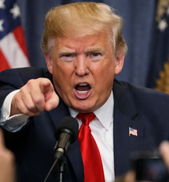 Donald Trump señalando de forma enérgica hacia el frente durante una rueda de prensa, con gesto serio y boca abierta mientras habla ante los micrófonos, con banderas de Estados Unidos desenfocadas al fondo y periodistas en primer plano.