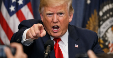 Donald Trump señalando de forma enérgica hacia el frente durante una rueda de prensa, con gesto serio y boca abierta mientras habla ante los micrófonos, con banderas de Estados Unidos desenfocadas al fondo y periodistas en primer plano.