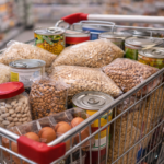 Fotografía en primer plano de un carrito de metal lleno hasta arriba con productos básicos de alimentación dentro de un supermercado. Se observan de forma clara bolsas transparentes de legumbres secas, arroz y pasta, varias latas de conserva apiladas, docenas de huevos en sus envases y botes de cristal. La imagen, con un enfoque nítido en los productos y el pasillo del supermercado desenfocado al fondo, transmite una sensación de compra de despensa completa y económica.