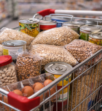 Fotografía en primer plano de un carrito de metal lleno hasta arriba con productos básicos de alimentación dentro de un supermercado. Se observan de forma clara bolsas transparentes de legumbres secas, arroz y pasta, varias latas de conserva apiladas, docenas de huevos en sus envases y botes de cristal. La imagen, con un enfoque nítido en los productos y el pasillo del supermercado desenfocado al fondo, transmite una sensación de compra de despensa completa y económica.